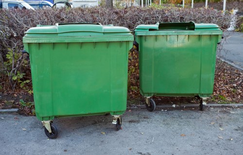Two large green plastic wheelie bins with black wheels are positioned side by side on a paved surface in an outdoor setting. The bins are rectangular with slightly curved lids, and one appears slightly taller than the other. They are situated in front of low, dense shrubbery and bare trees, with a metal fence visible in the background. The surface beneath the bins is a grey pavement or driveway, with some scattered fallen leaves nearby. The scene is illuminated by natural daylight, giving a neutral and clear view of the bins and their surroundings, consistent with typical rubbish removal or waste collection areas often found in residential or commercial premises within Mitcham or nearby areas in south London.