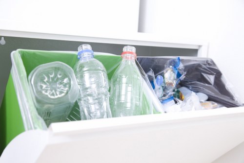 The image shows a close-up view of a white open drawer or bin containing two clear plastic bottles in the foreground, with one featuring a blue cap and the other a red cap, both positioned horizontally. Behind the bottles, there is a black plastic bag filled with mixed waste, possibly packaging or miscellaneous rubbish, with part of the bag folded over. To the left, a green plastic container or recycling bin is partially visible, holding a transparent plastic container with a lid. The background appears to be an indoor setting with a white surface, possibly a countertop or other storage area, suggesting a location associated with household or office rubbish collection. The scene emphasizes waste disposal and recycling materials, aligned with rubbish removal services such as those provided by Office Clearance Mitcham, serving the Mitcham area nearby CR4 postcode.