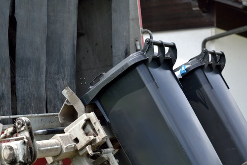 The image shows a close-up of two black wheelie bins positioned on a metal frame, likely attached to a waste collection vehicle. The bins are made of durable plastic with a smooth, matte finish, and their lids are securely closed. One bin has a blue plastic bottle visible near the opening. The surrounding environment features a grey metallic surface in the background, possibly part of the waste vehicle or nearby structure, with a faint hint of a wooden or dark panel surface above. The bins are set on a paved area, indicating an outdoor setting typical for rubbish collection or waste disposal in a residential or commercial area. This scene visually supports rubbish removal services provided by Office Clearance Mitcham, illustrating the equipment used for waste collection in Mitcham or nearby areas within south London.