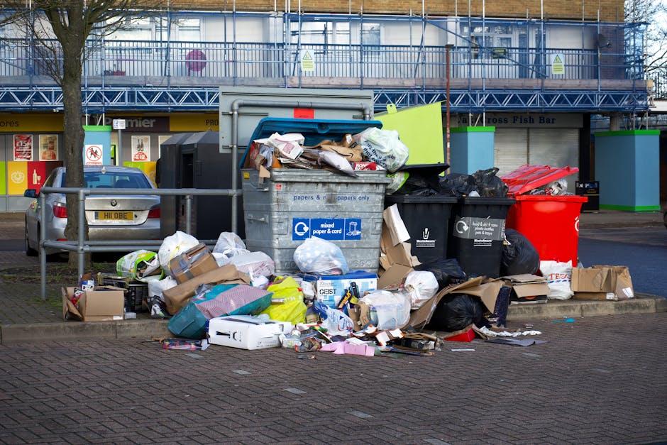 A waste collection operative dressed in green high-visibility workwear with reflective orange stripes stands beside a rear-loading rubbish collection vehicle on a tree-lined urban street. The vehicle,