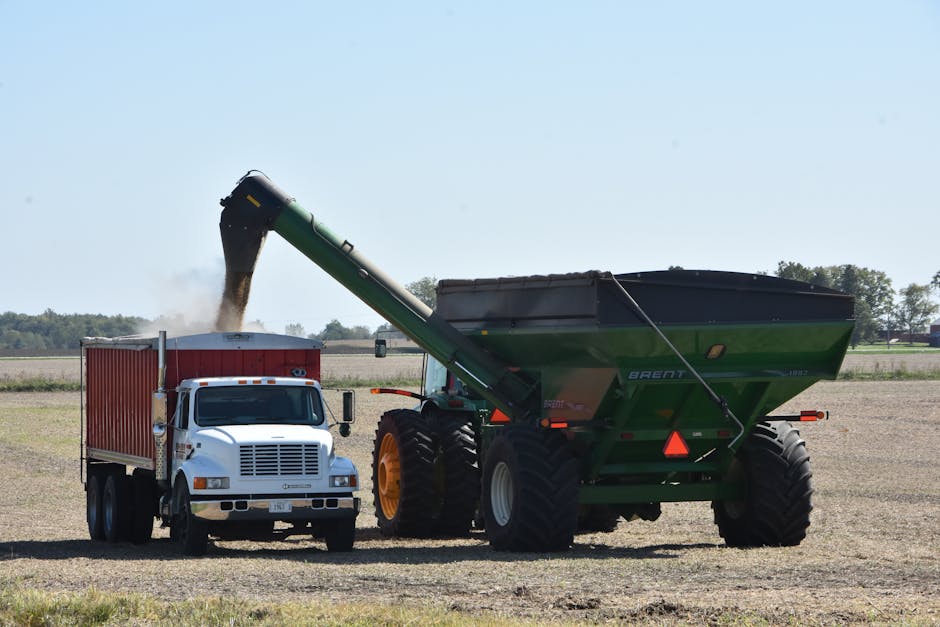 A large green and black agricultural tractor with oversized tires is positioned in a flat, open field, actively unloading a dark, granular material from its long conveyor chute into a nearby white and