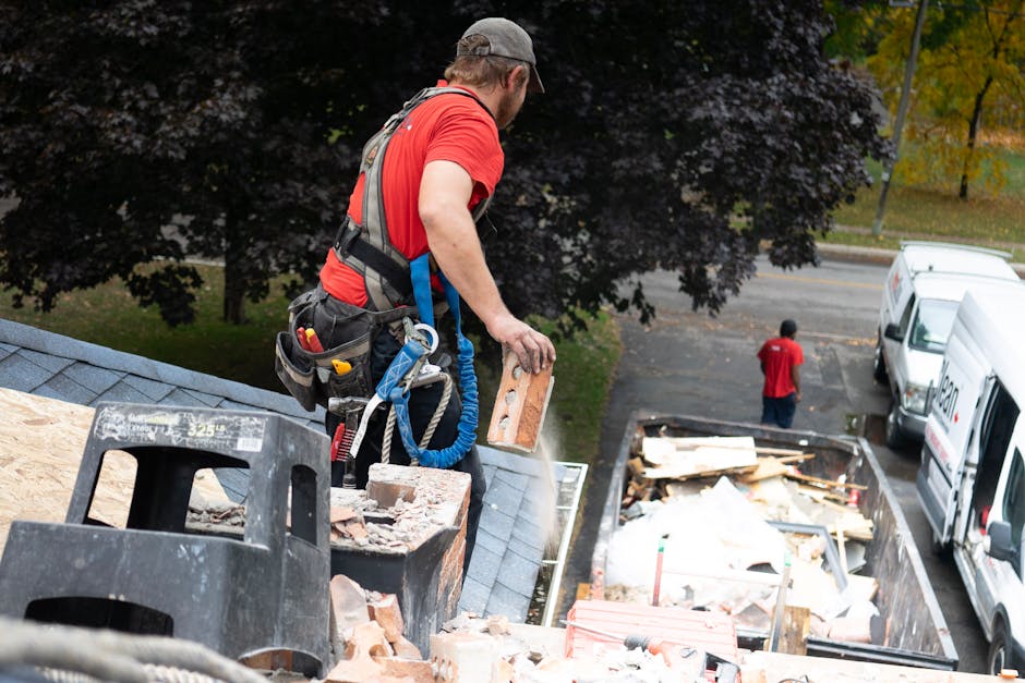 A compact yellow and black excavator with a hydraulic arm and bucket is actively demolishing a section of a white residential building's exterior wall, which has a textured finish. The scene is set ou
