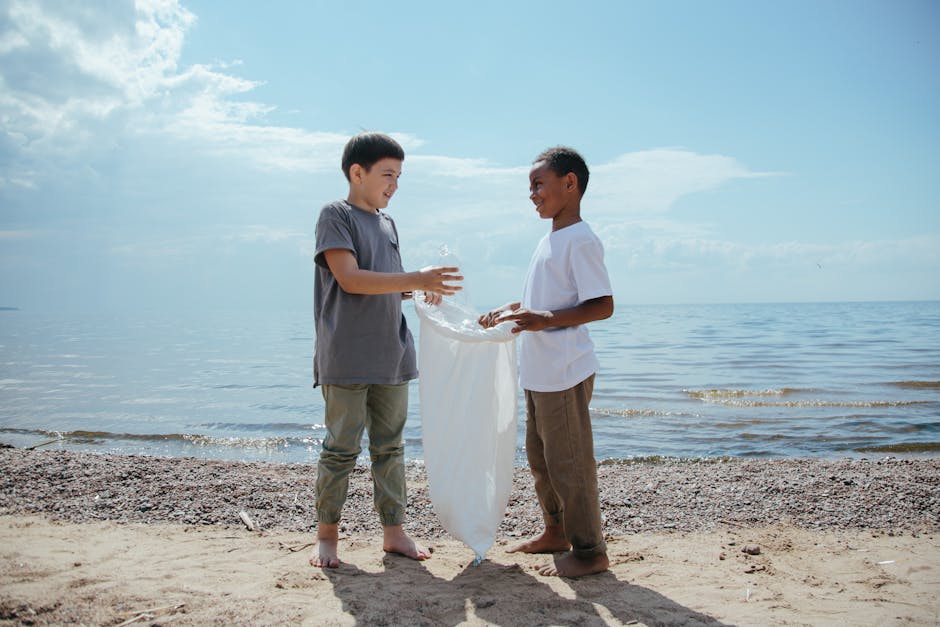 Two young boys stand on a sandy beach near the water's edge under a partly cloudy sky. One boy, dressed in a grey t-shirt and light grey pants, holds a large, clear plastic bag filled with water, whil